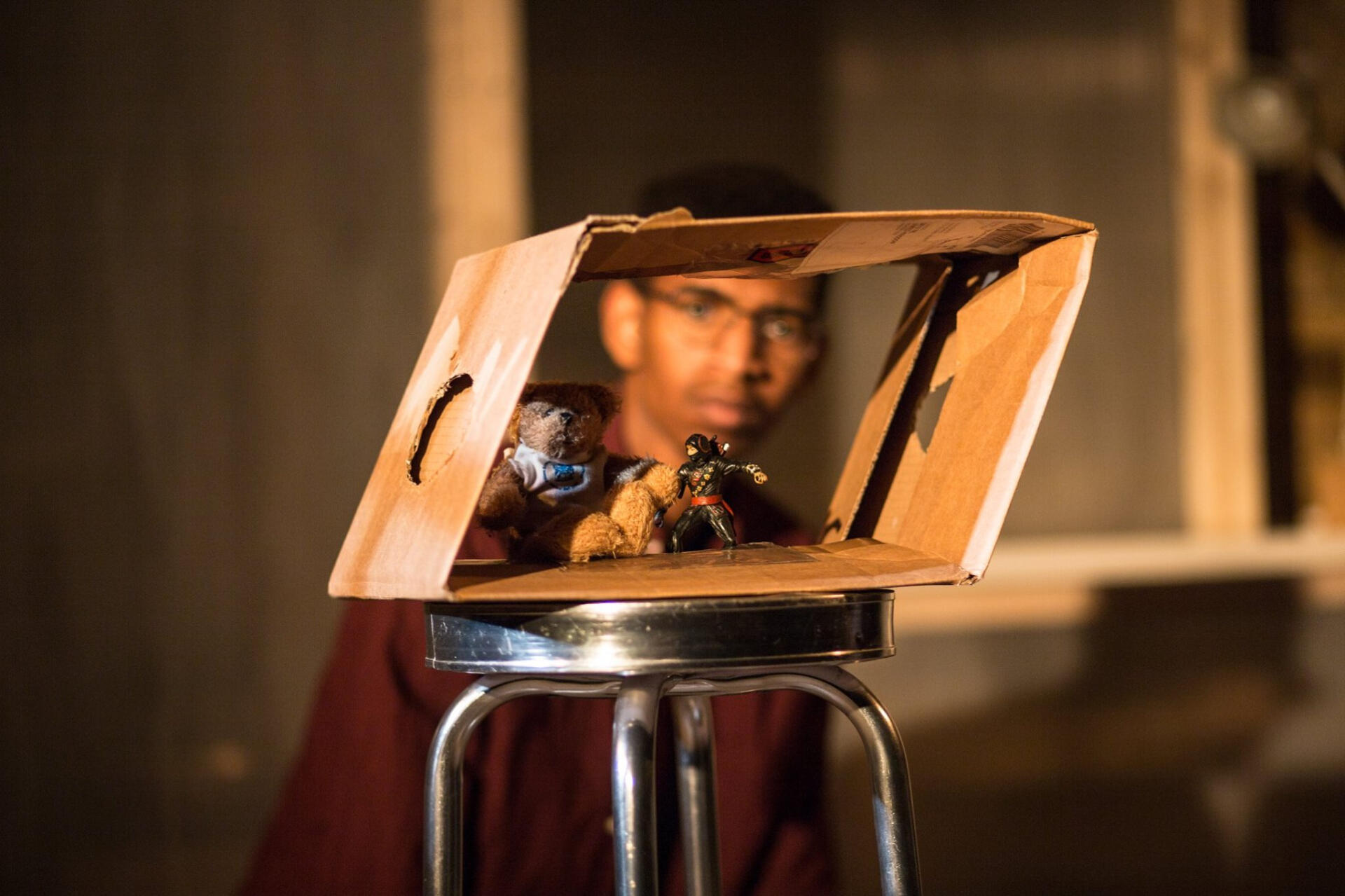 A teddy bear and a ninja action figure in a box watched by a man